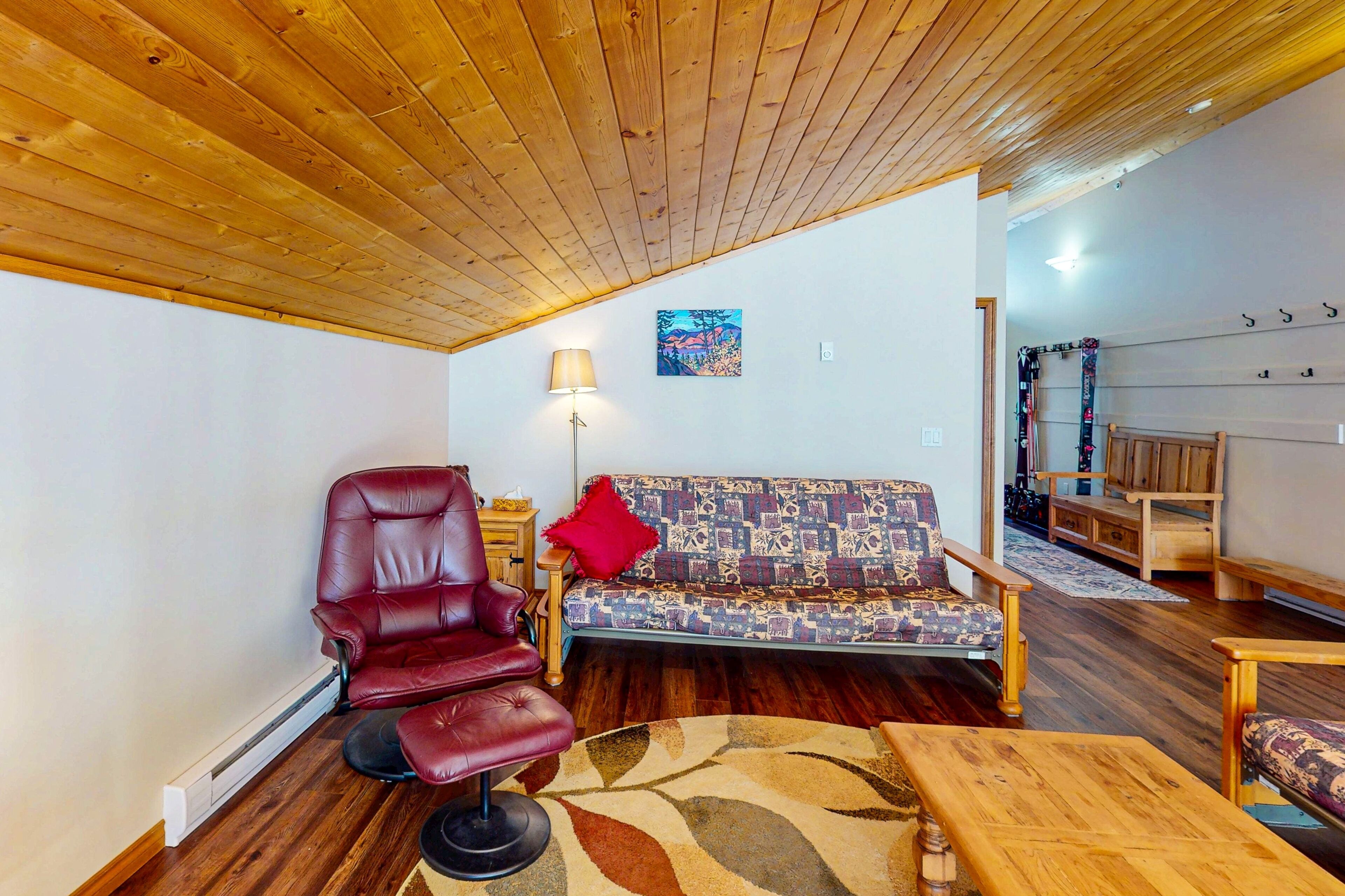 A living room with a wooden ceiling, a red leather chair, a patterned sofa, a lamp, and a wooden table.