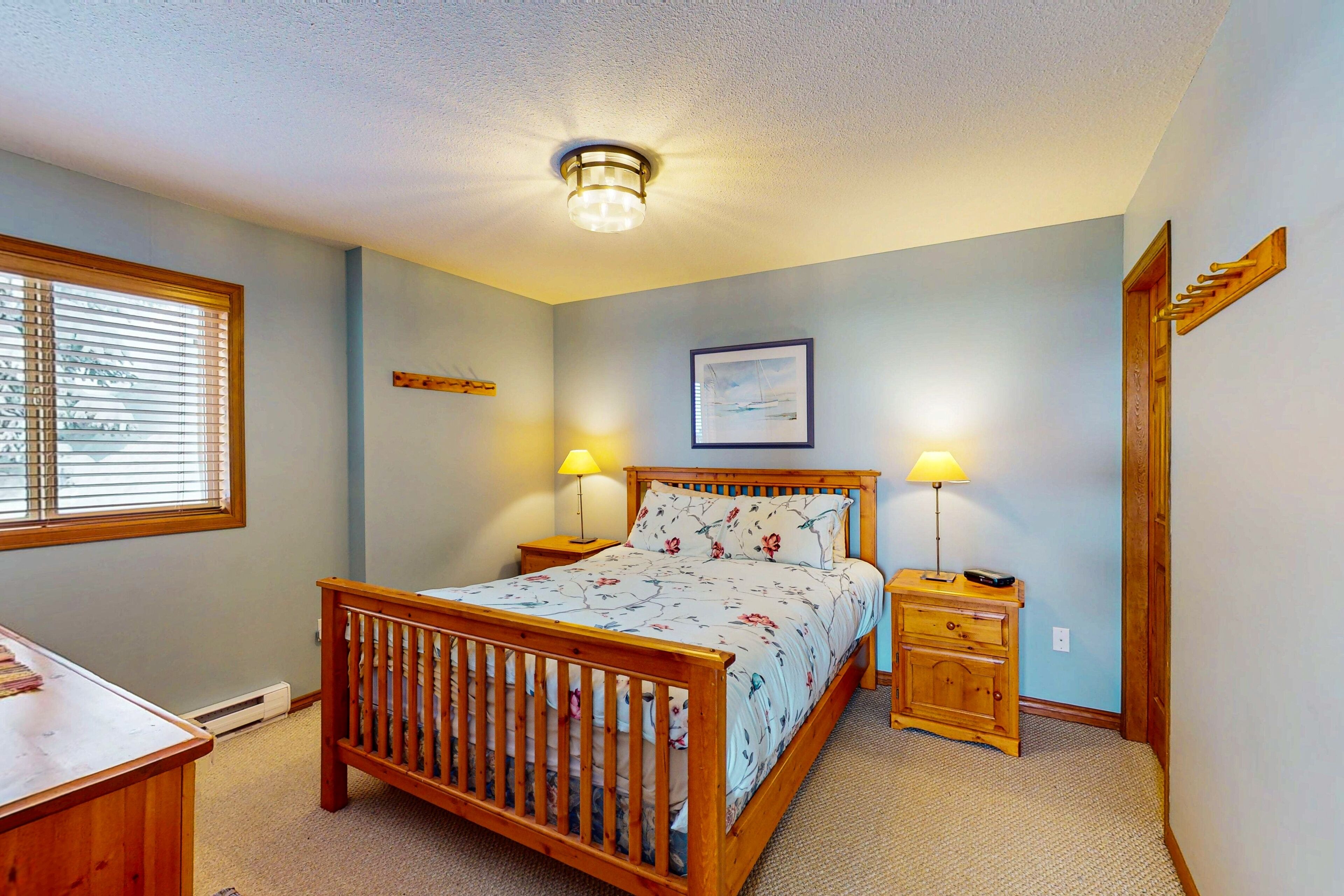 A bedroom with a wooden bed, two bedside tables with lamps, a window with blinds, a framed picture on the wall, and a wooden door with a coat rack.