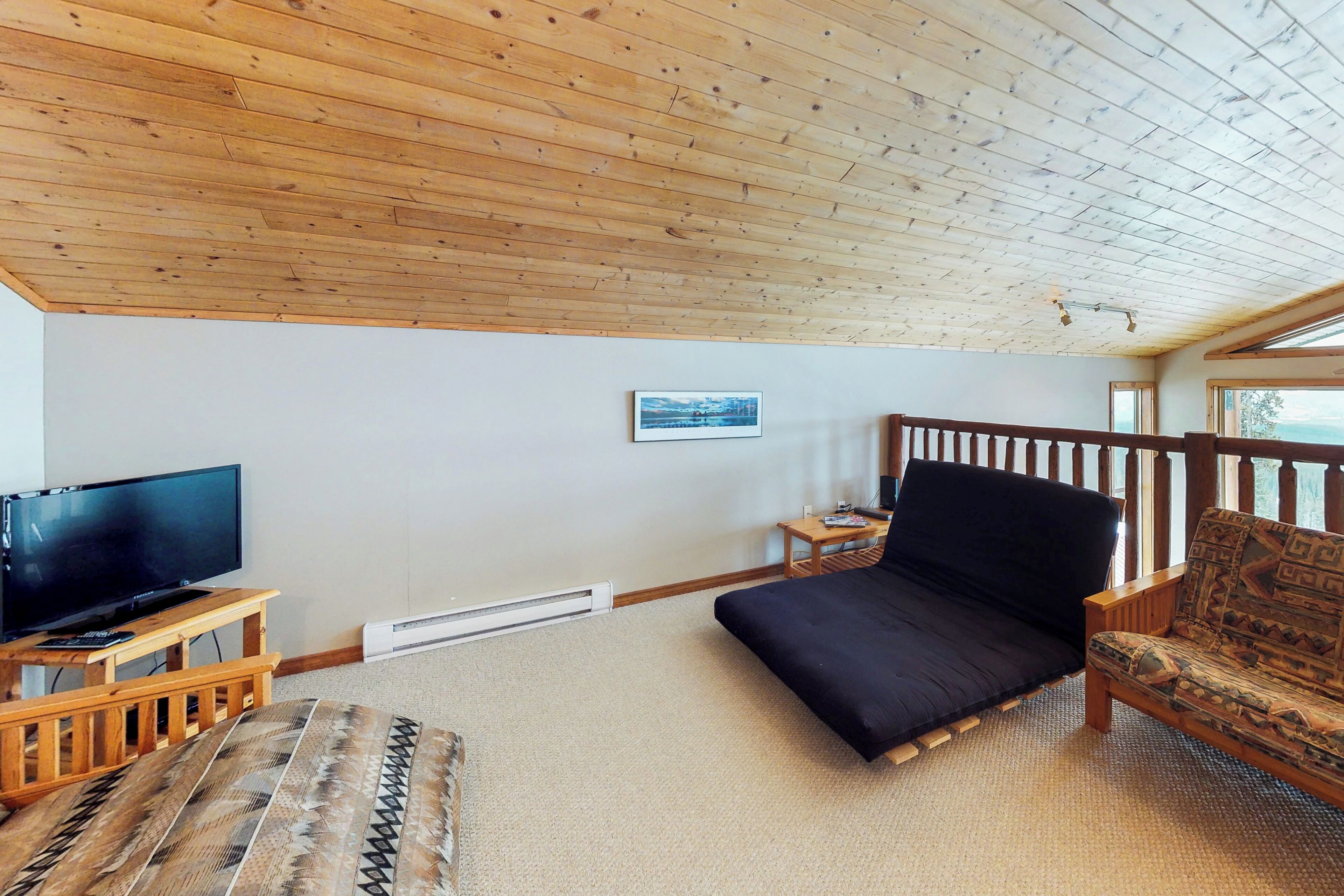 A cozy room with a wooden ceiling, a flat-screen TV on a wooden stand, a dark sofa, a patterned ottoman, and a small table with a chair.
