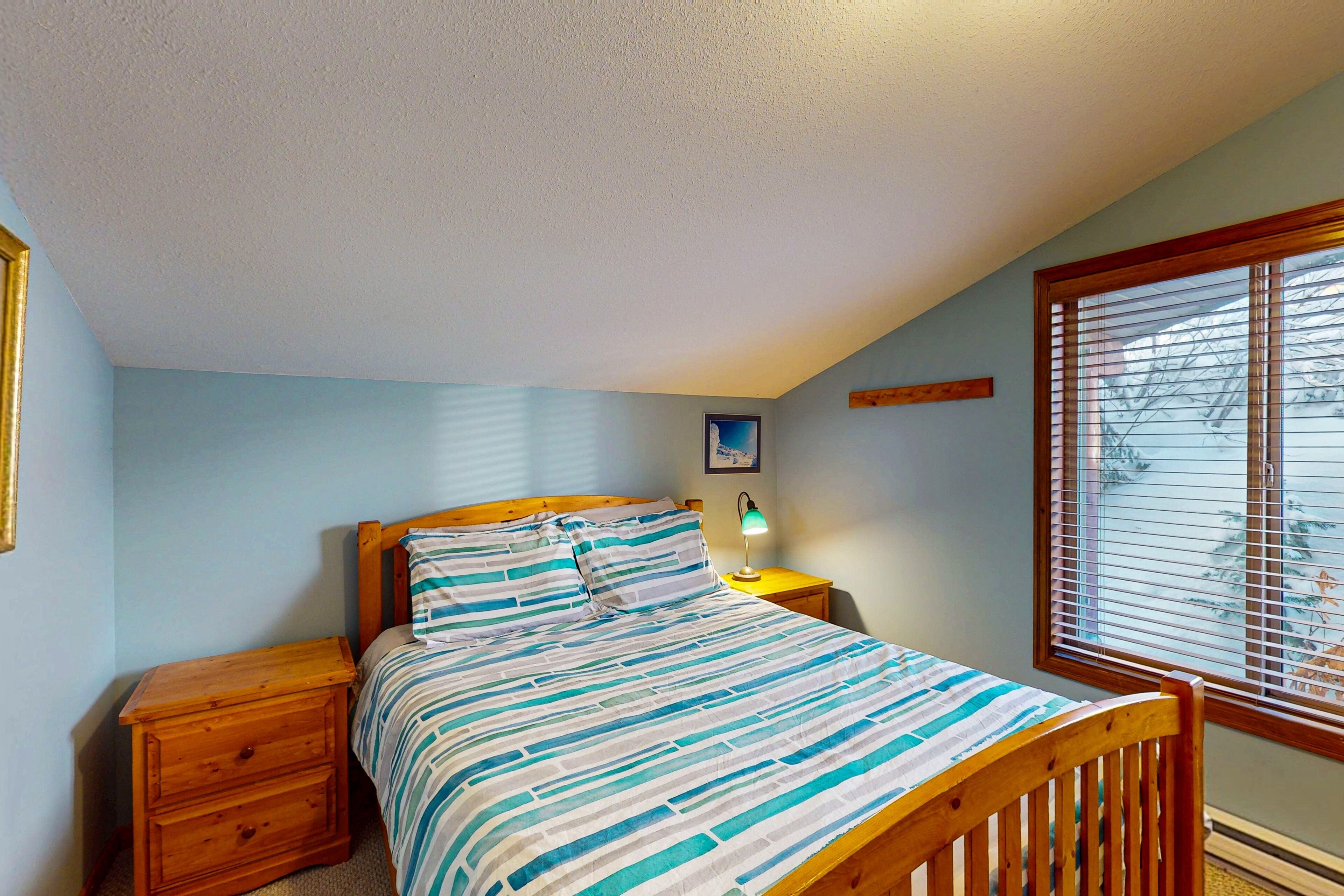A bedroom with a bed, wooden nightstands, a night lamp, a window with blinds, and a picture on the wall.