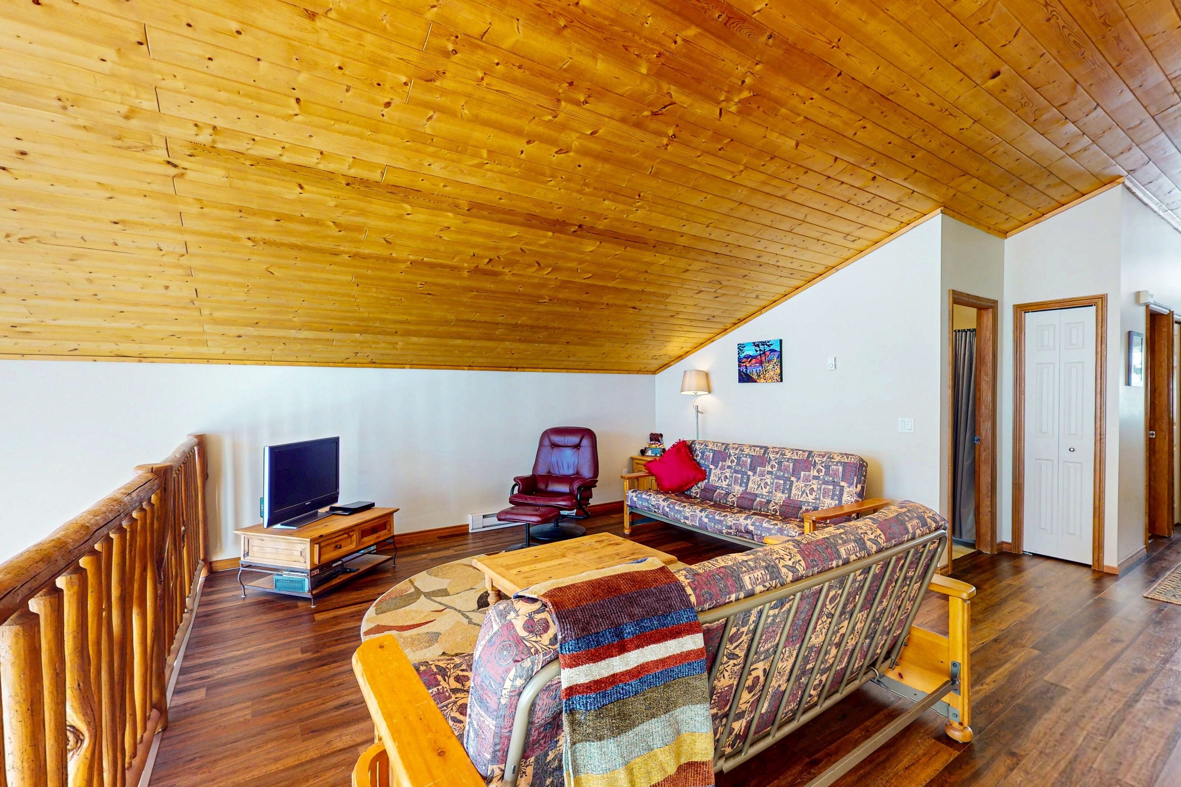 A room with a wooden ceiling, a sofa, a chair, a television, and a wooden floor.