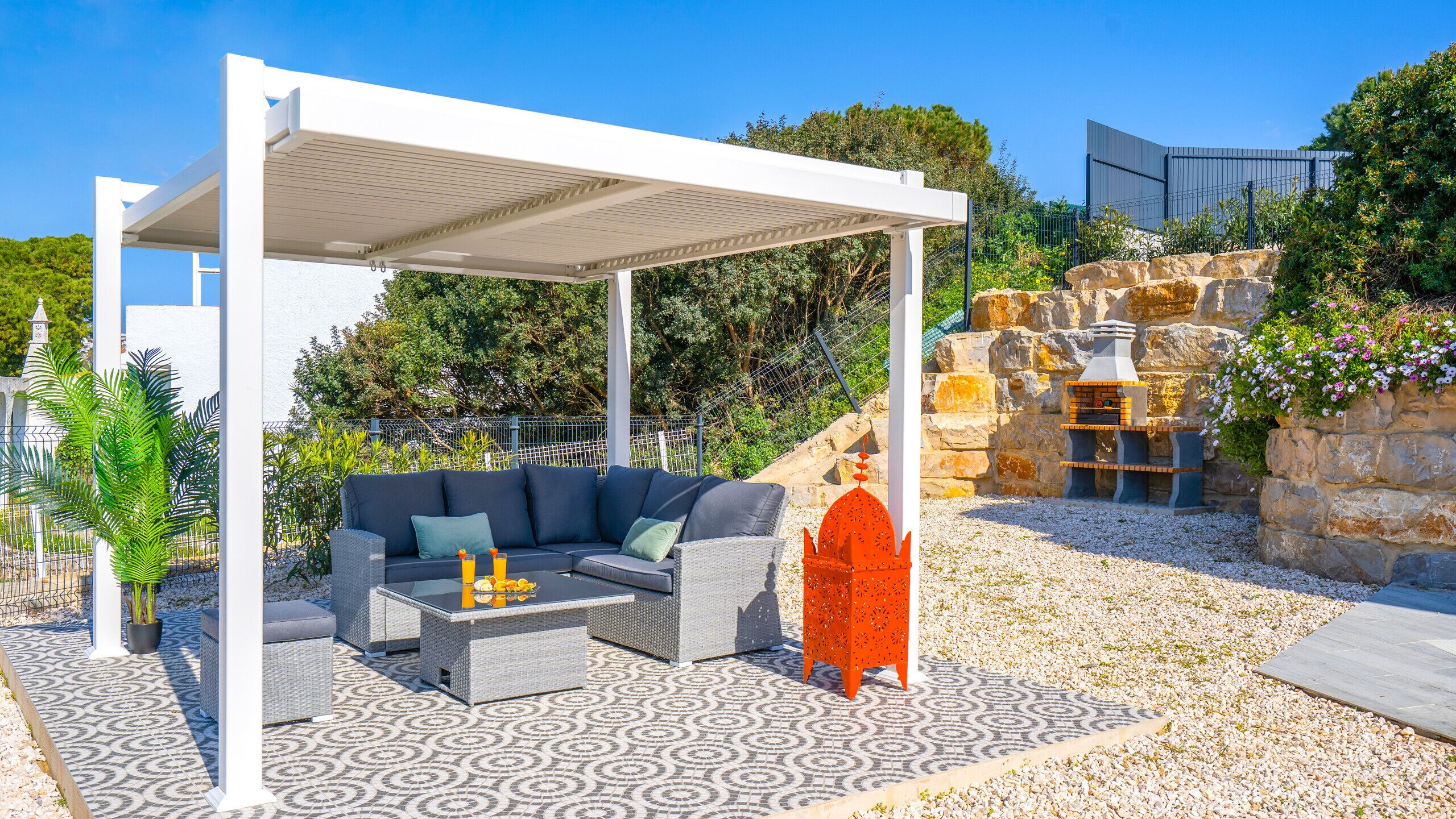 A covered outdoor seating area with a sofa and table, a decorative orange structure, and a stone wall with a pizza oven.