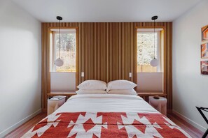 Quarto com cama, duas mesas de cabeceira, tapete vermelho estampado, paredes de ripas de madeira e duas janelas com persianas.