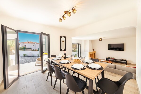 Un comedor moderno con una mesa de madera dispuesta para seis personas, rodeada de sillas, y un amplio ventanal que ofrece vistas a un edificio blanco y follaje exterior.
