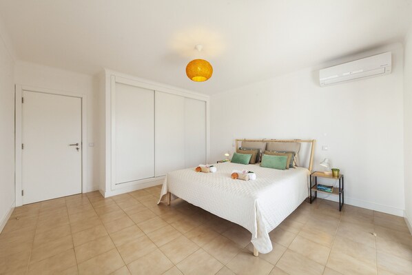 Un dormitorio con cama, paredes blancas, una lámpara de techo, aire acondicionado y una mesita de noche con lámpara.