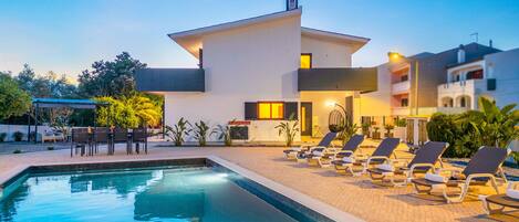 A modern hotel with a swimming pool, sun loungers, and a covered outdoor seating area.
