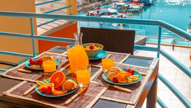 Una mesa de desayuno con fruta, jugo y pasteles en un balcón con vista a un puerto deportivo.