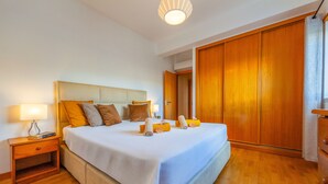Un lit bien fait, recouvert d’une couvre-lit blanche et bleue, une table de chevet en bois avec une lampe, et une armoire en bois dans une pièce bien éclairée.
