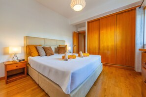 A neatly made bed with a white and blue bedspread, a wooden nightstand with a lamp, and a wooden wardrobe in a well-lit room.