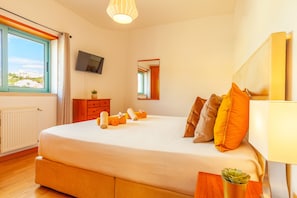 Un dormitorio con una cama grande, un televisor en la pared, una ventana con cortinas y una planta pequeña en la mesita de noche.