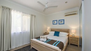 A hotel room with a bed, two bedside tables, a ceiling fan, an air conditioning unit, and a window with curtains.
