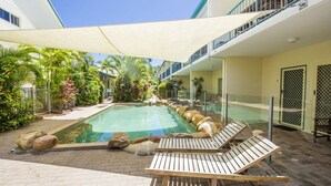 A pool area with lounge chairs, a shaded canopy, and a building with balconies.