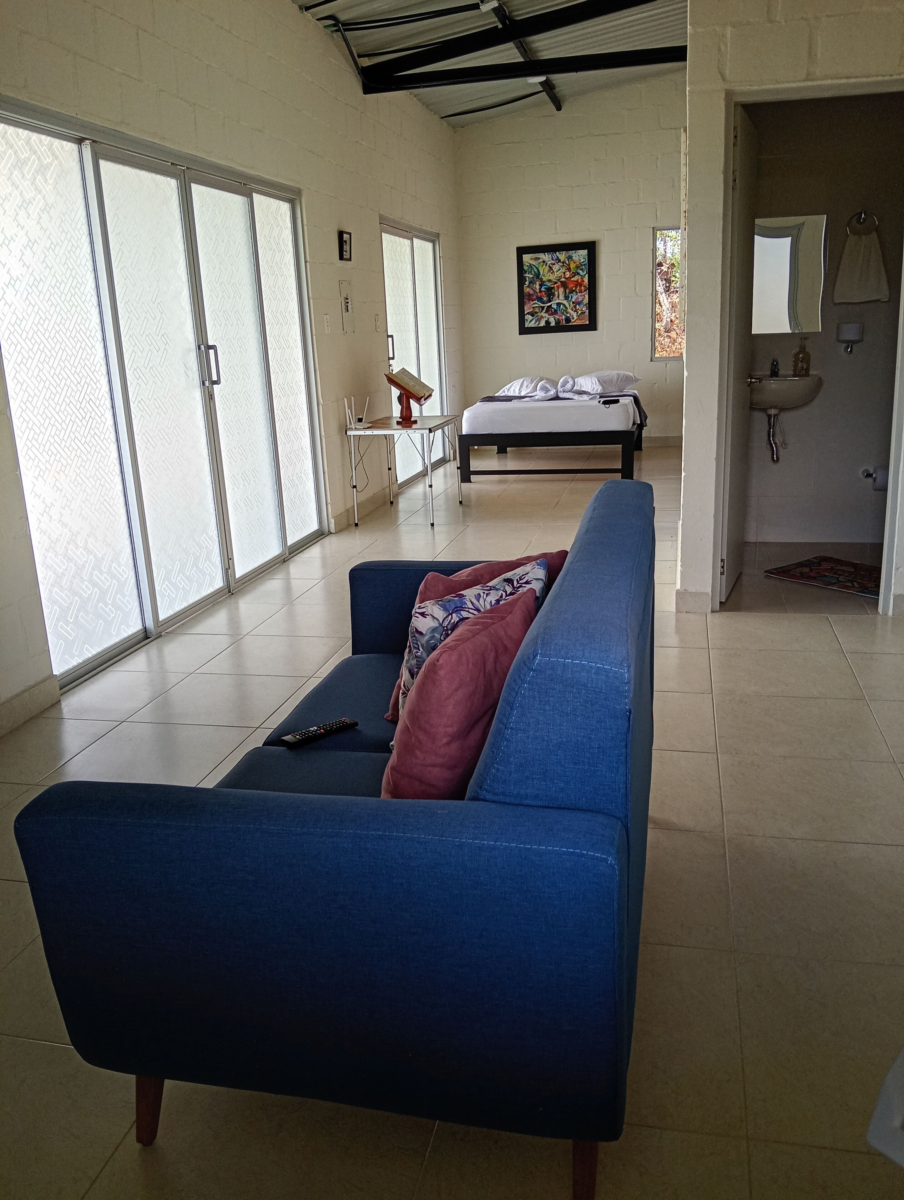 Une pièce avec un canapé bleu, un lit, un bureau avec une chaise, un tableau au mur et une salle de bain avec un lavabo et un miroir.