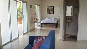 A room with a bed, a blue sofa, a small table, a painting, and a bathroom with a sink and mirror. - Macori Cabañas Campestres (Rivera)