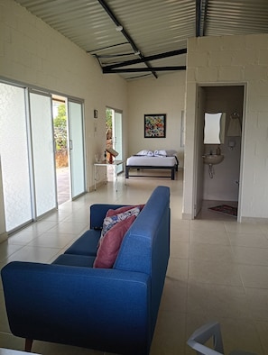 Una habitación con cama, sofá azul, mesita pequeña, un cuadro y baño con lavabo y espejo.