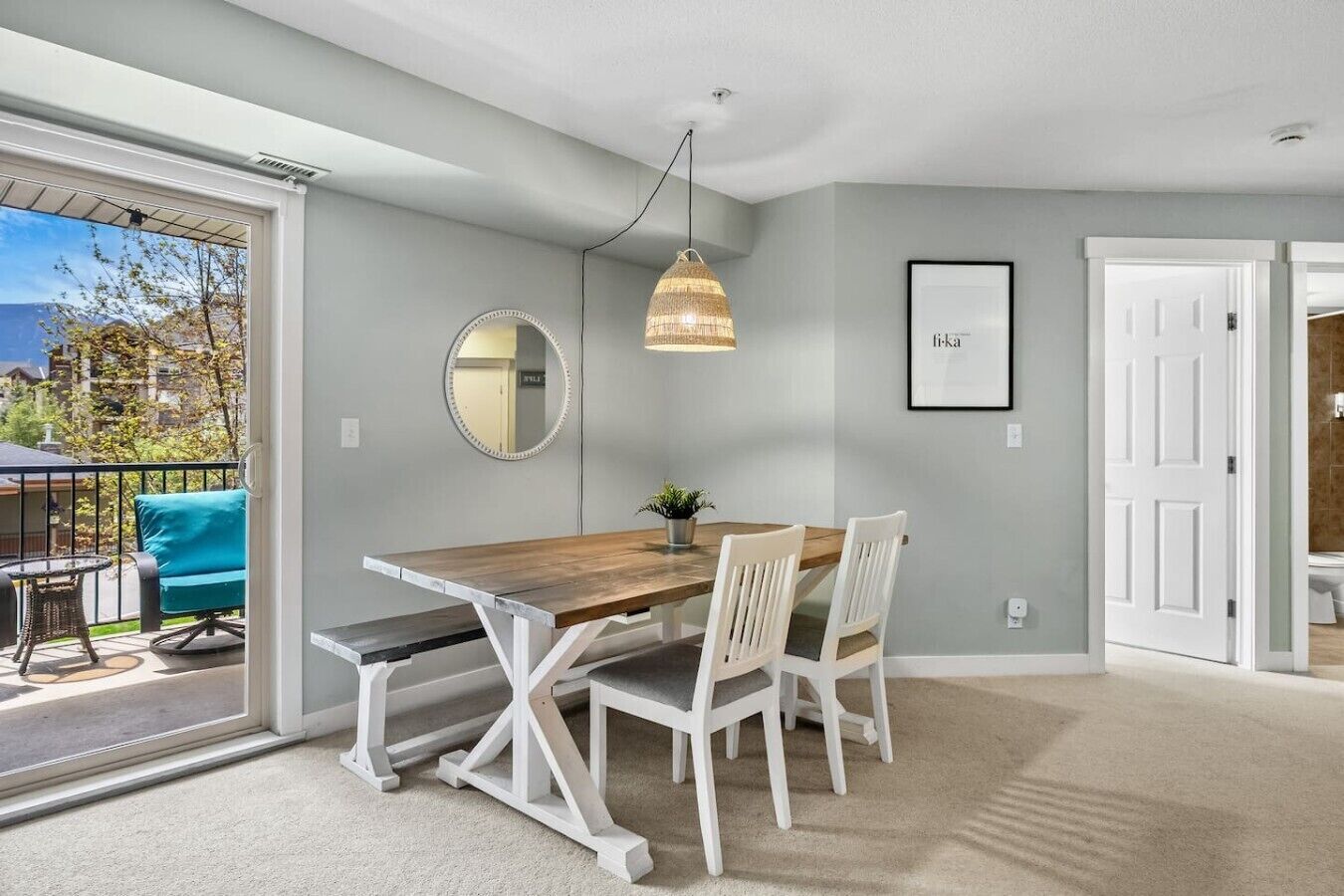 Un coin repas avec une table en bois, des chaises blanches, un luminaire suspendu, un miroir et un tableau encadré au mur.