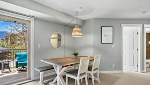 Un comedor con una mesa de madera, sillas blancas, una lámpara colgante, un espejo y un cuadro enmarcado en la pared.