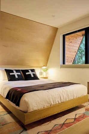 Ein Schlafzimmer mit Bett, Nachttisch, Fenster mit Blick auf Bäume und einer geneigten Decke.