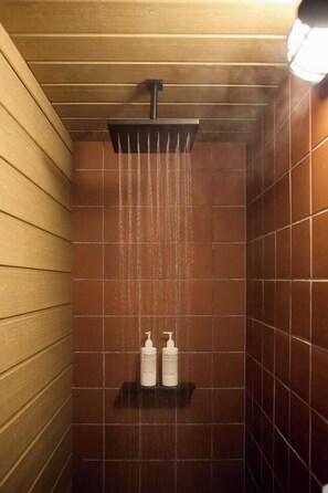 Eine Dusche mit Holzbalkendecke und braunen Fliesen, ein Handbrausekopf sowie zwei Flaschen auf einem Regal.
