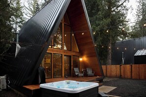 Eine moderne A-förmige Holzhütte mit Whirlpool, Holzterrasse und Lichterkette.
