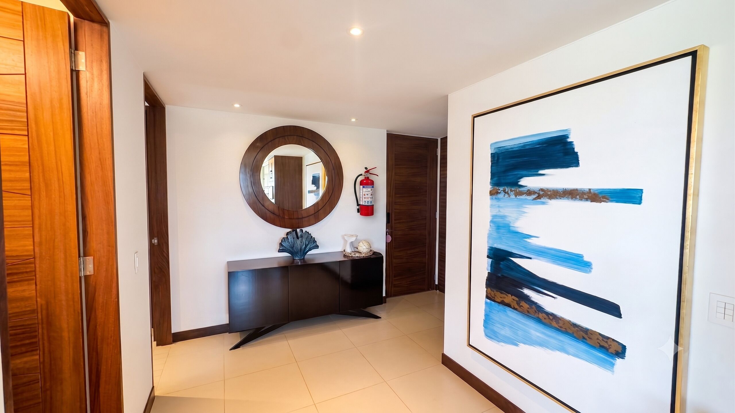 Een hal met een houten deur, een ronde spiegel, een donkere console en een schilderij met abstracte kunst, plus een brandblusser.