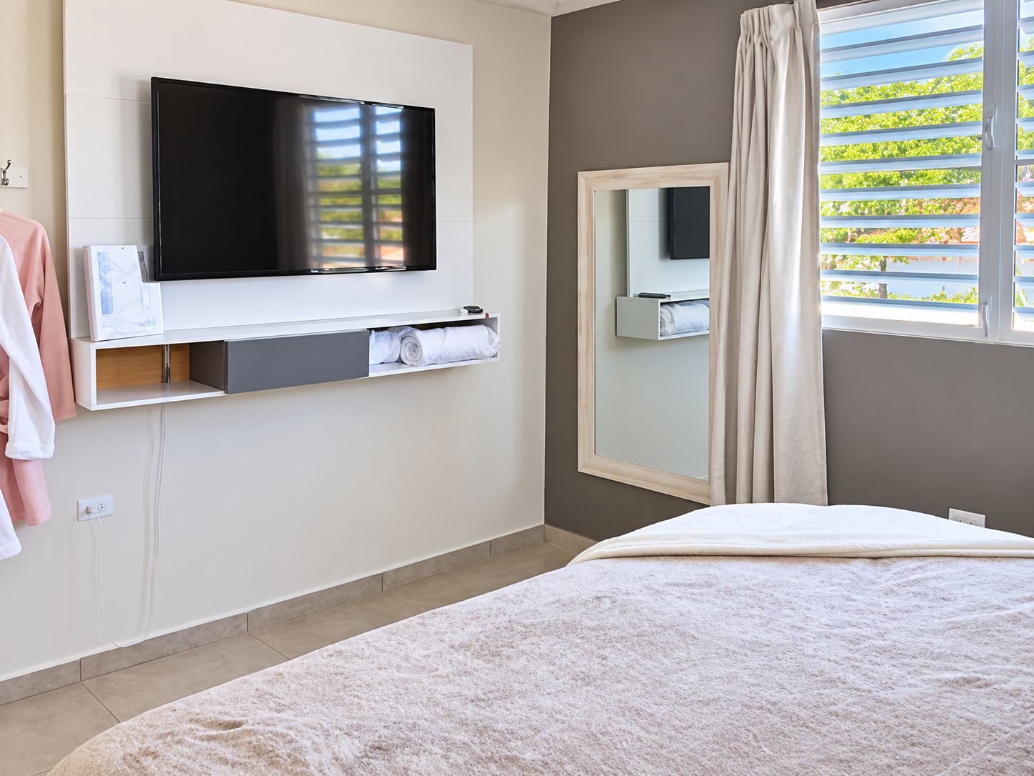 Habitación de hotel con televisor de pantalla plana montado en la pared, espejo, cama con edredón blanco y ventana con persianas.