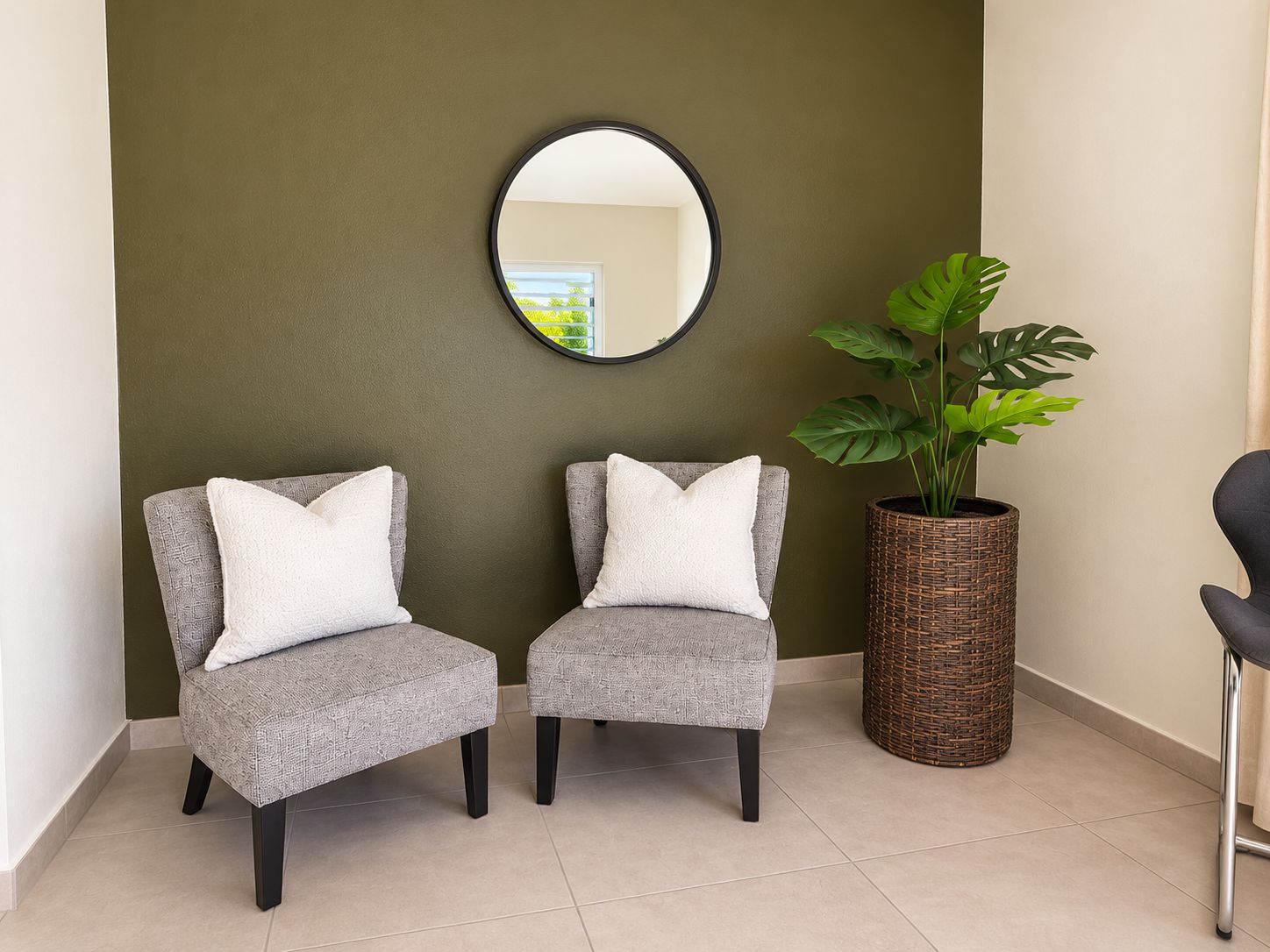 Two grey armchairs with white cushions, a round mirror on the wall, and a large potted plant with broad leaves.