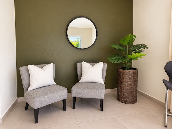 Two grey armchairs with white cushions, a round mirror on the wall, and a large potted plant with broad leaves.