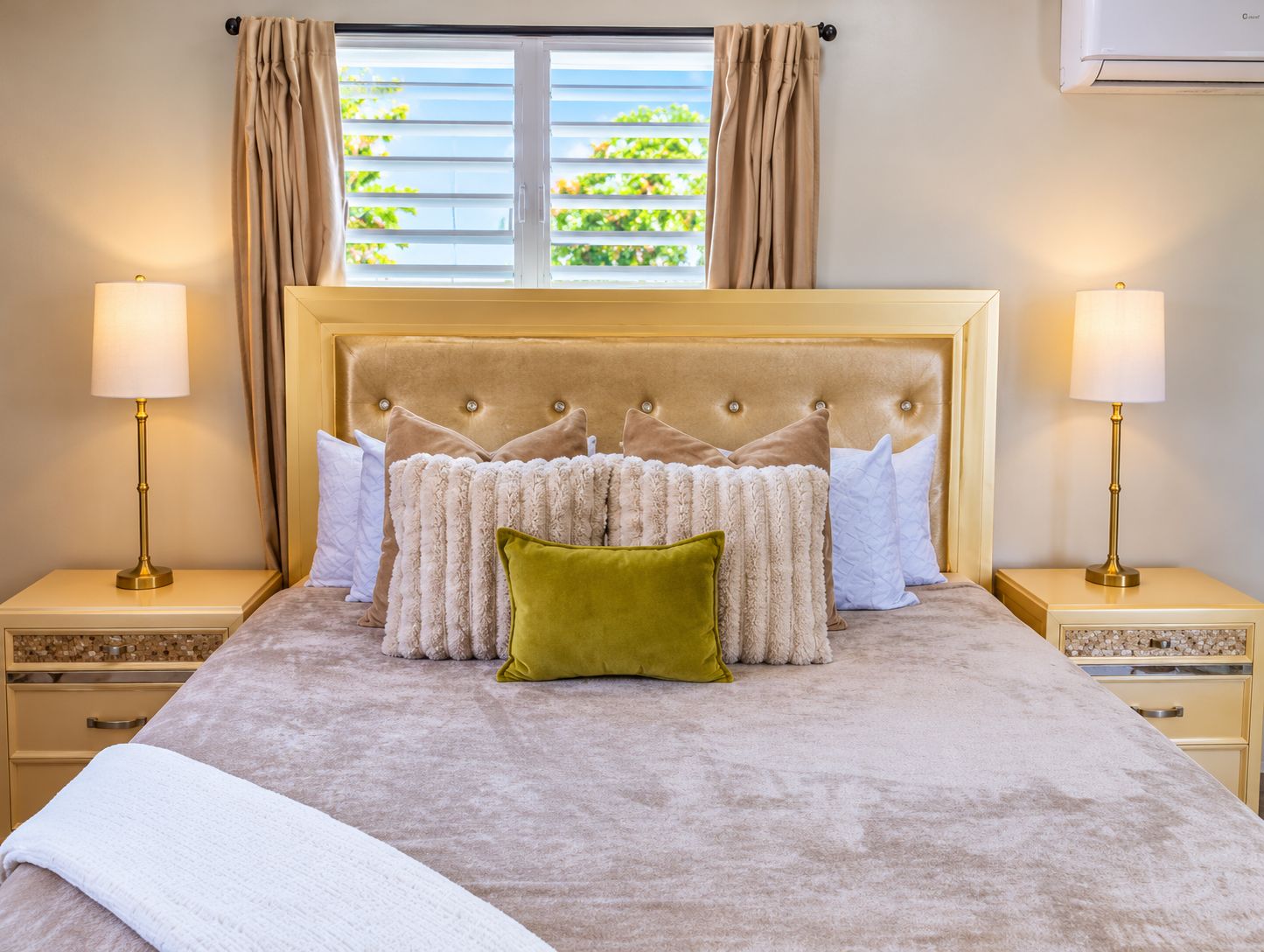 A bedroom with a large bed, two bedside tables with lamps, a window with curtains, and an air conditioning unit.