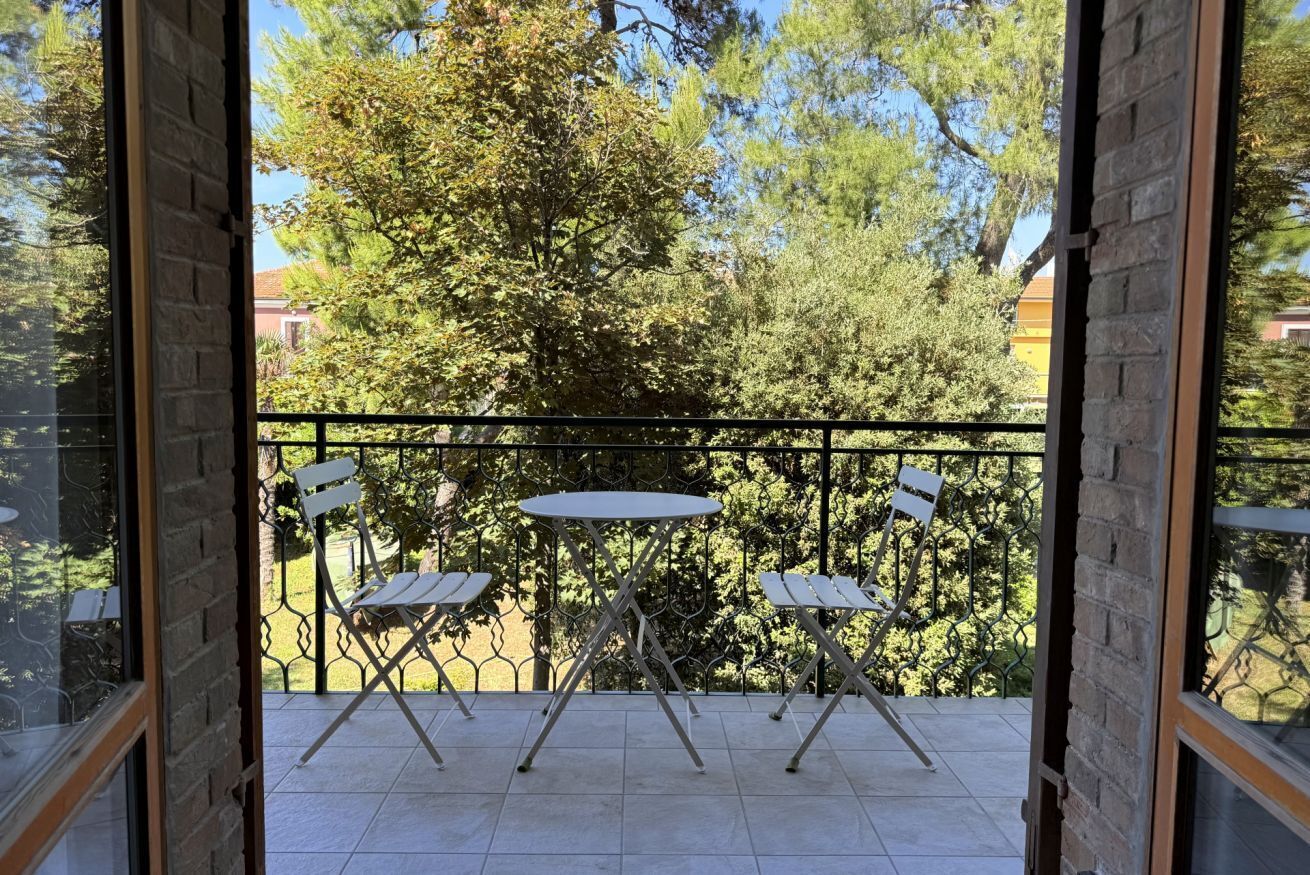 An open balcony with a table and chairs, a metal railing, and a view of trees and buildings.