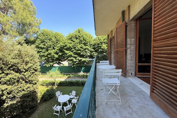 A balcony with white metal chairs and tables, a swimming pool, and a view of trees and a building.