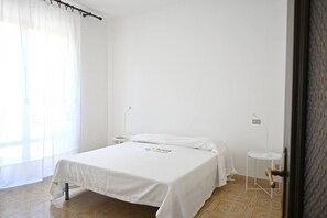 Ein helles, minimalistisches Hotelzimmer mit einem Bett, einem kleinen Nachttisch, einer Lampe und einem Fenster mit durchsichtigen Vorhängen.