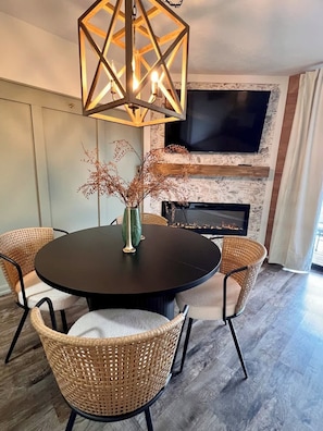 A round dining table with a dark surface, surrounded by wicker chairs with black metal legs. Above the table, there is a geometric pendant light fixture. - Our Nest-Pointe Royale Resort-Pools/Hot Tub/Golf (Branson)