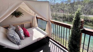 Un balcon doté d’un espace salon avec des coussins, d’un auvent blanc et offrant une vue sur une rivière et des arbres.
