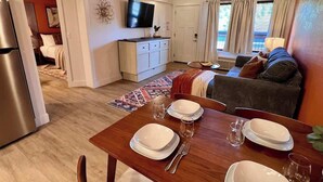 Ein Essbereich mit einem Holztisch, gedeckt für vier Personen, eine Küche mit Kühlschrank, Fernseher und einem Sofa im Hintergrund.