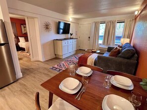Ein Essbereich mit einem Holztisch, gedeckt für vier Personen, eine Küche mit Kühlschrank, Fernseher und einem Sofa im Hintergrund.
