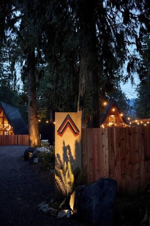 Una valla de madera con un cartel triangular, una cabaña de madera y un árbol grande.