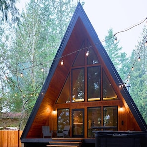 Eine dreieckige Holzhütte mit großen Fenstern, einer Veranda mit Sitzgelegenheiten und Lichterketten.