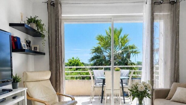 Ein modernes Wohnzimmer mit Fernseher, einer Couch, einem Sessel, einem Couchtisch und einem Balkon mit Gartenmöbeln.