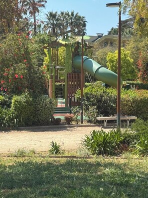 Ein Spielplatz mit Rutsche, umgeben von Grünflächen und einer Bank.
