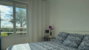Ein Schlafzimmer mit einem Bett, einem Nachttisch, einer Lampe und einem Fenster mit Blick auf Palmen.
