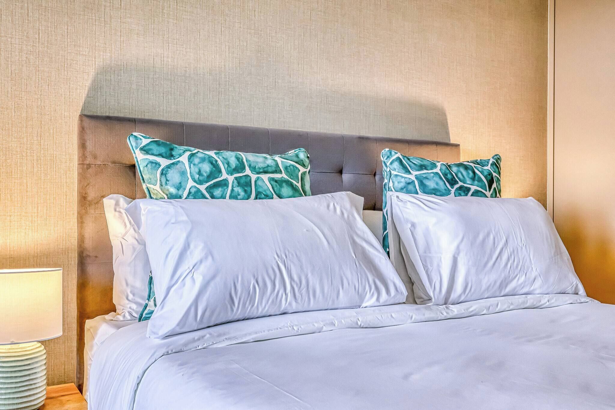 Una cama con ropa de cama blanca y dos almohadas estampadas en verde, una lámpara de noche y una pared con textura.