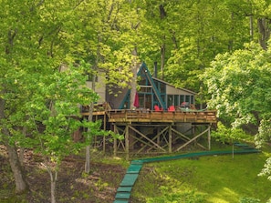 A wooden deck with red chairs, a house with a green roof, and a green pathway leading to a water feature.