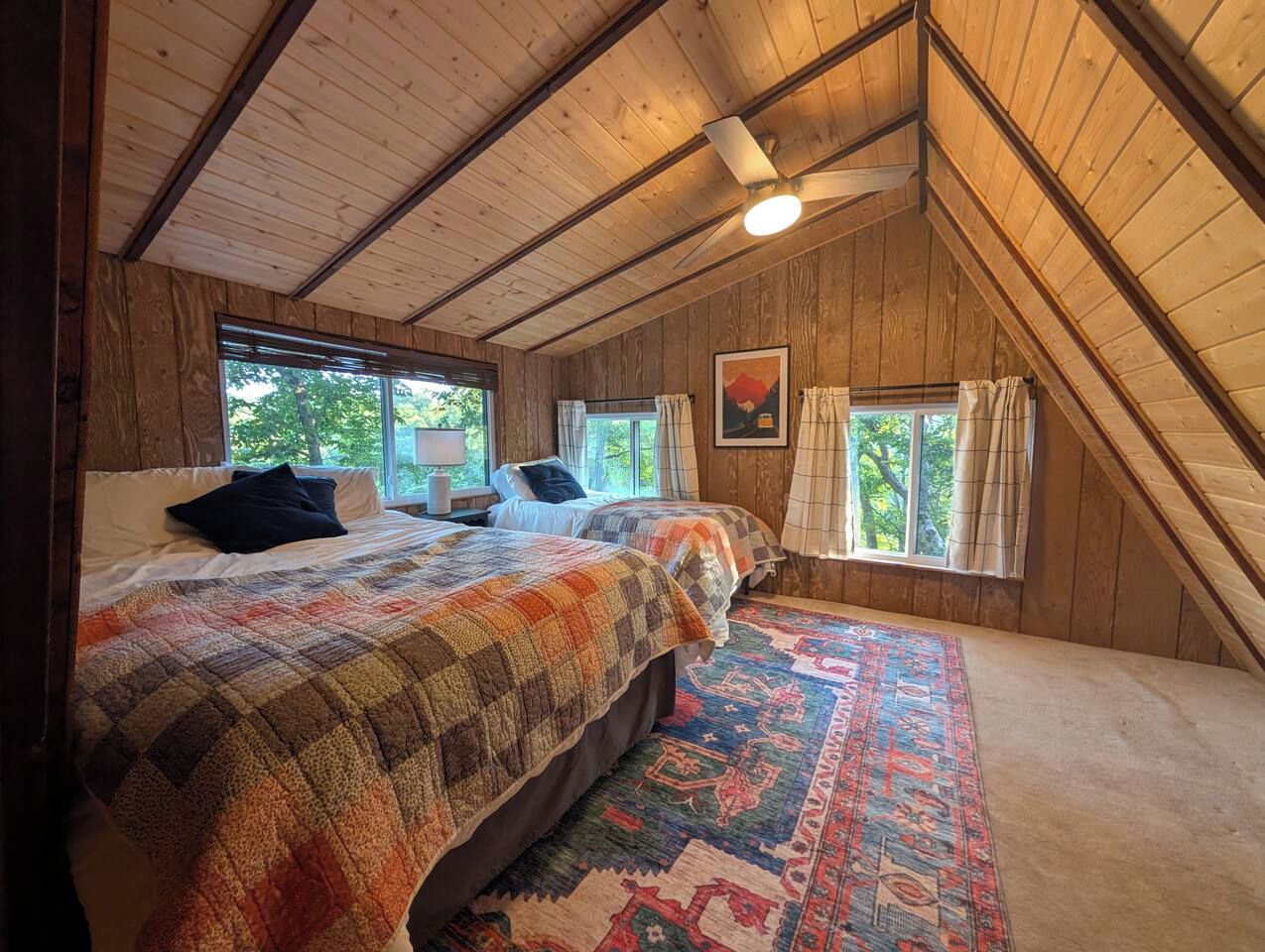 Ein Schlafzimmer mit schrägem Dach, zwei Betten, einem großen Fenster und einem farbenfrohen Teppich.