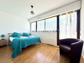 A bedroom with a bed, a chair, a nightstand, a lamp, and a window with curtains. - T2 Thermes et centre Aix-les-Bains parking (Aix-les-Bains)