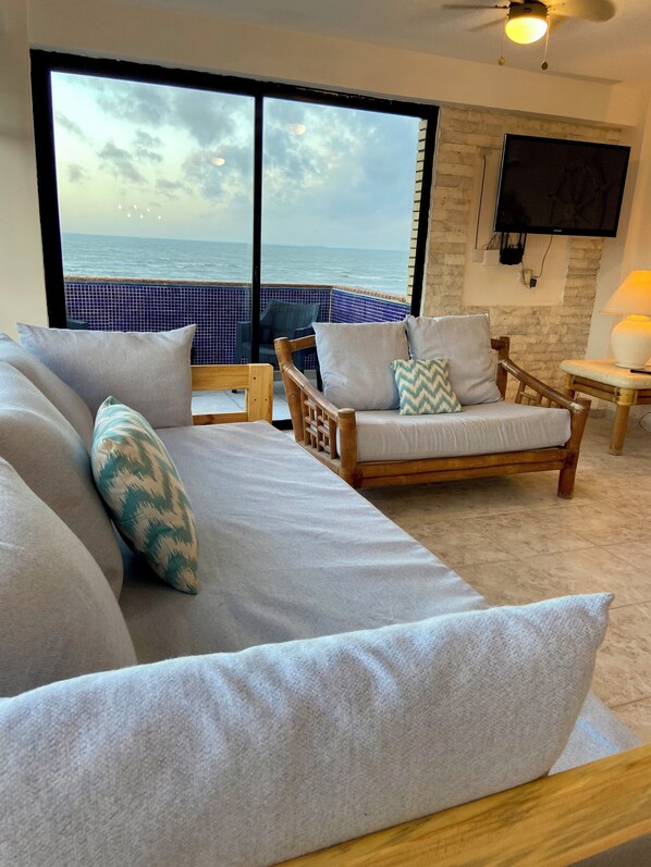 Una habitación con un ventanal grande con vistas al mar, un sofá con cojines, una silla de madera y una mesita con una lámpara.