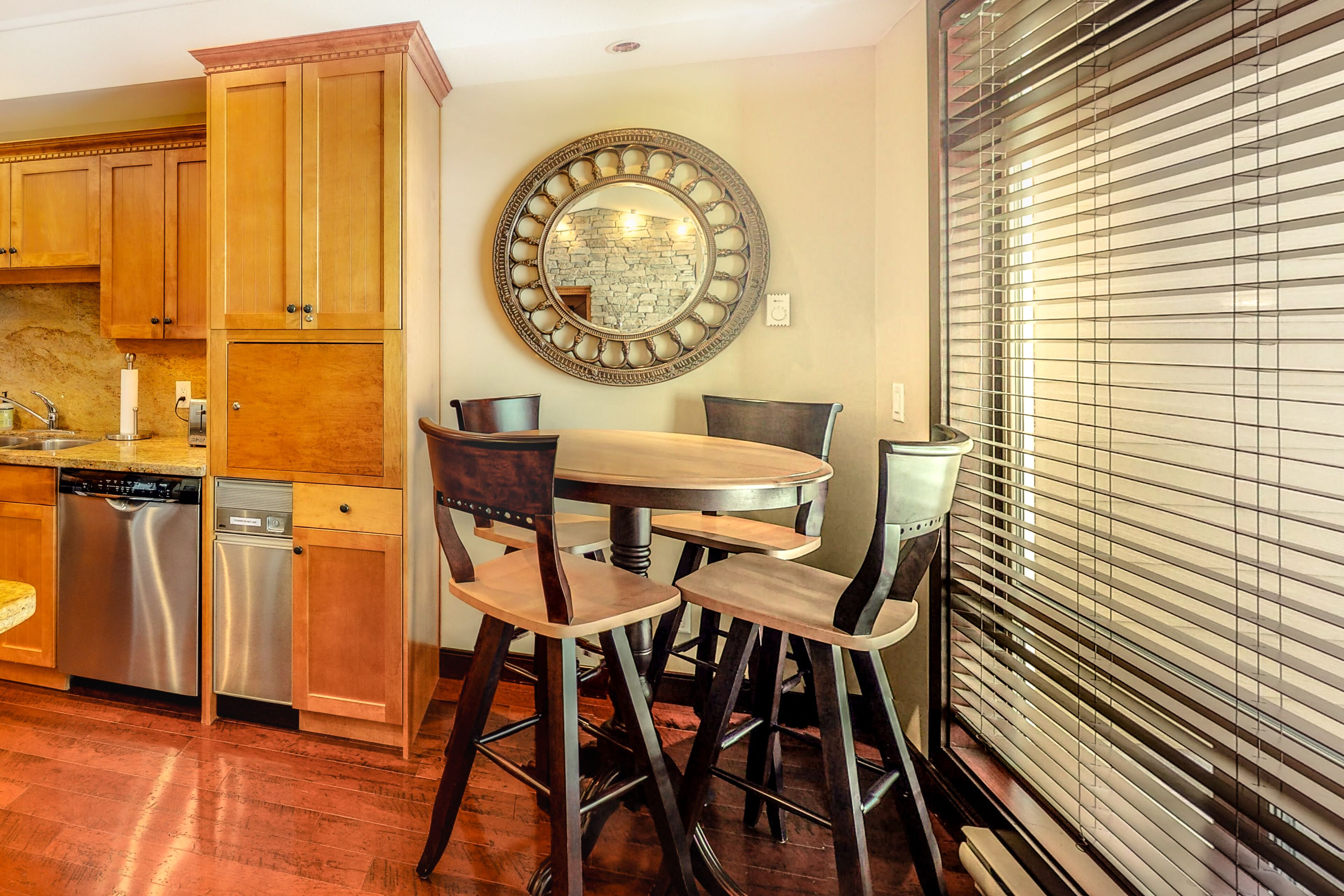 A kitchen with wooden cabinets, a stainless steel dishwasher, a round mirror, and a dining area with stools.