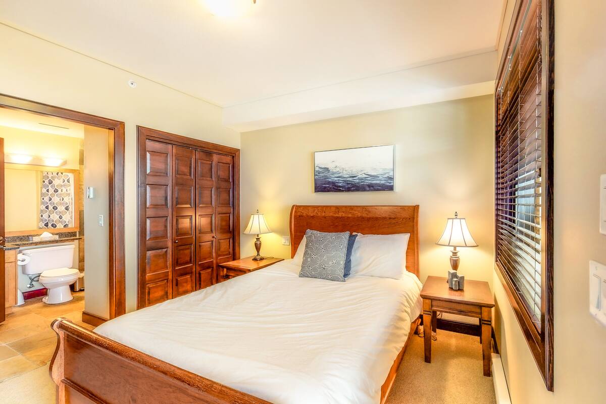 A hotel room with a large bed, wooden headboard, two bedside tables with lamps, a bathroom with a toilet and sink, and a window with blinds.