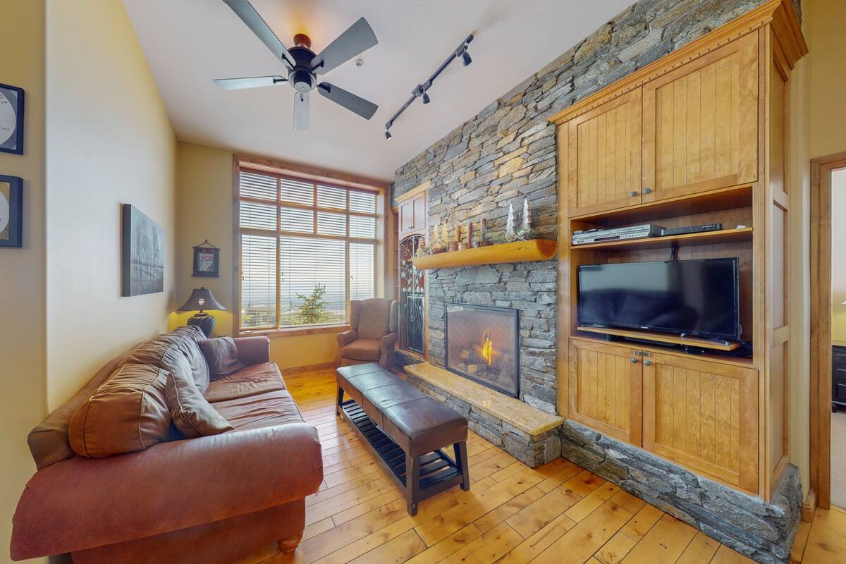 A cozy living room with a stone fireplace, wooden shelves, a leather sofa, and a flat-screen TV.
