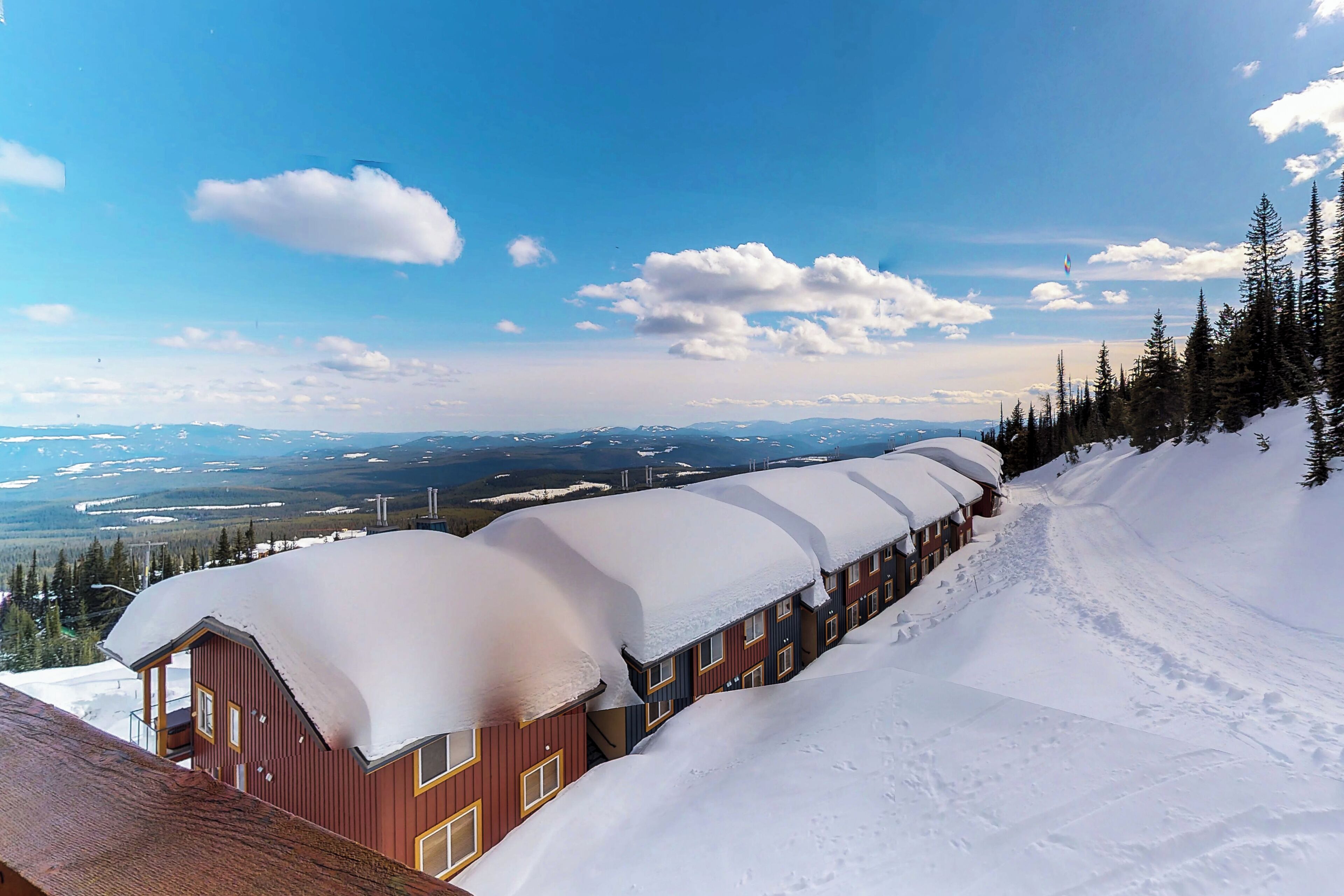 A row of cabins with snow-covered roofs, surrounded by snow-covered ground and trees, with a clear sky in the background.